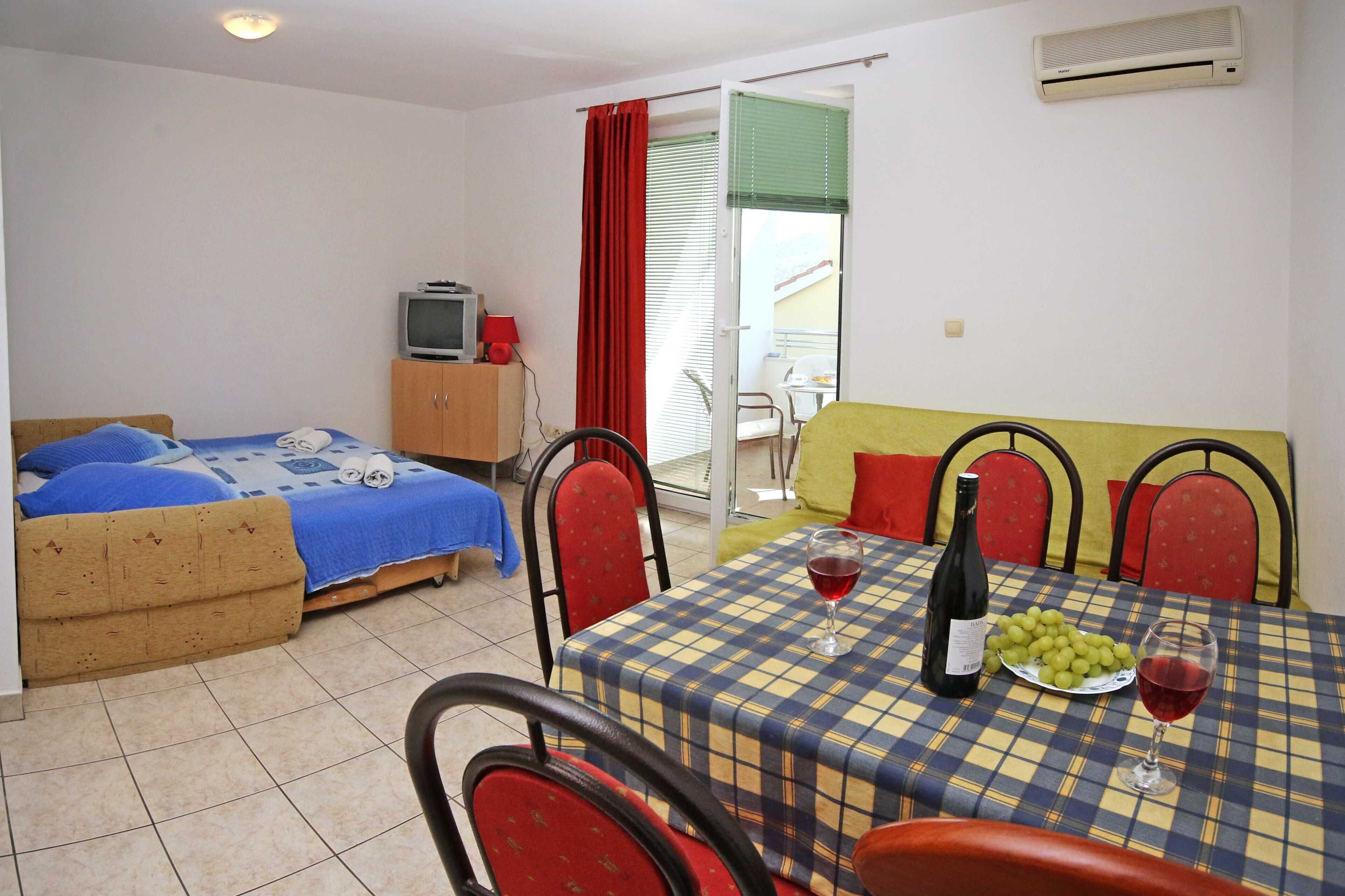 Ein Zimmer mit Bett, kleinem Fernseher, Esstisch mit Stühlen und einem Balkon mit Sitzgelegenheit.