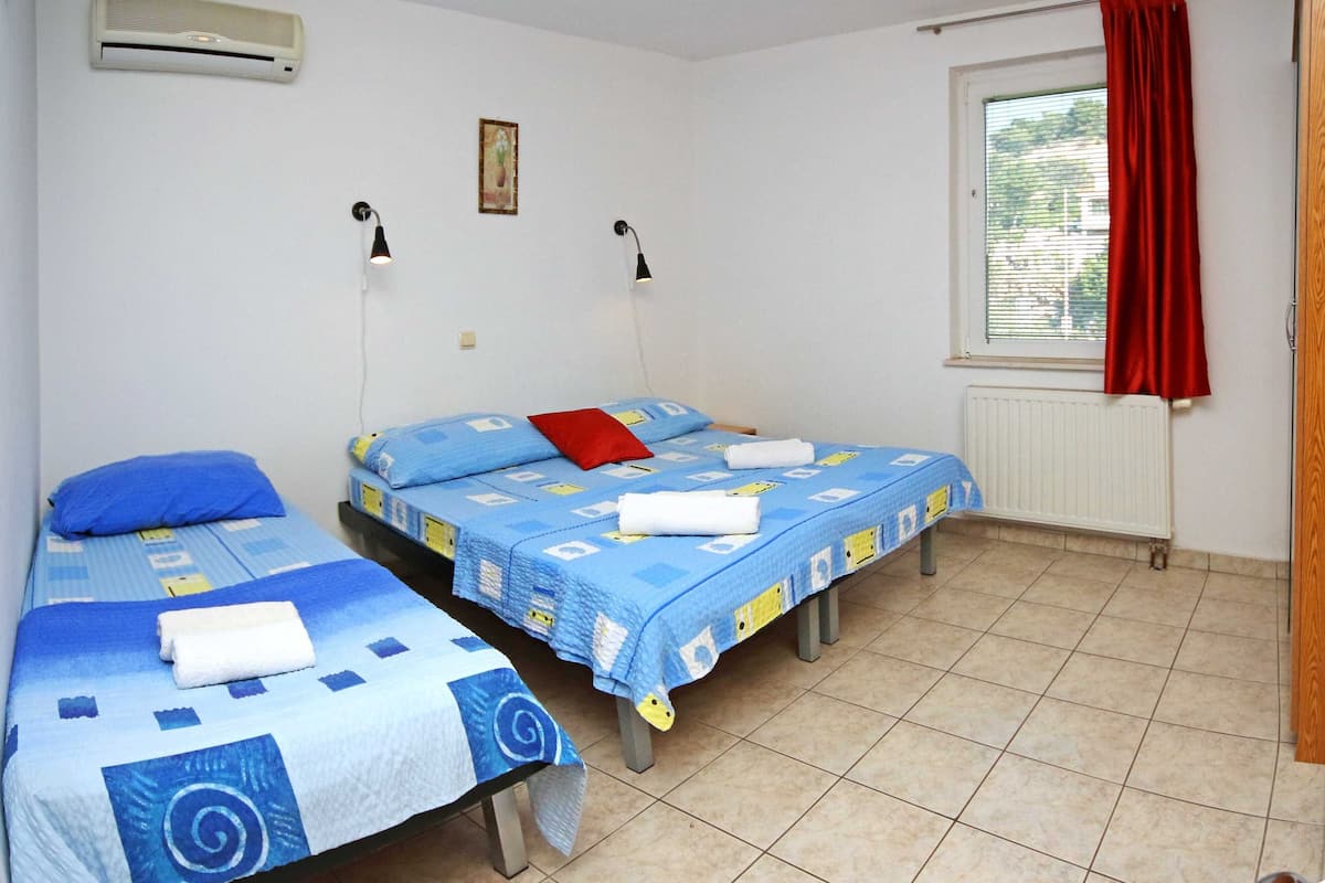 A room with two beds, each with blue bedding and a red pillow, a window with red curtains, and a radiator on the wall.