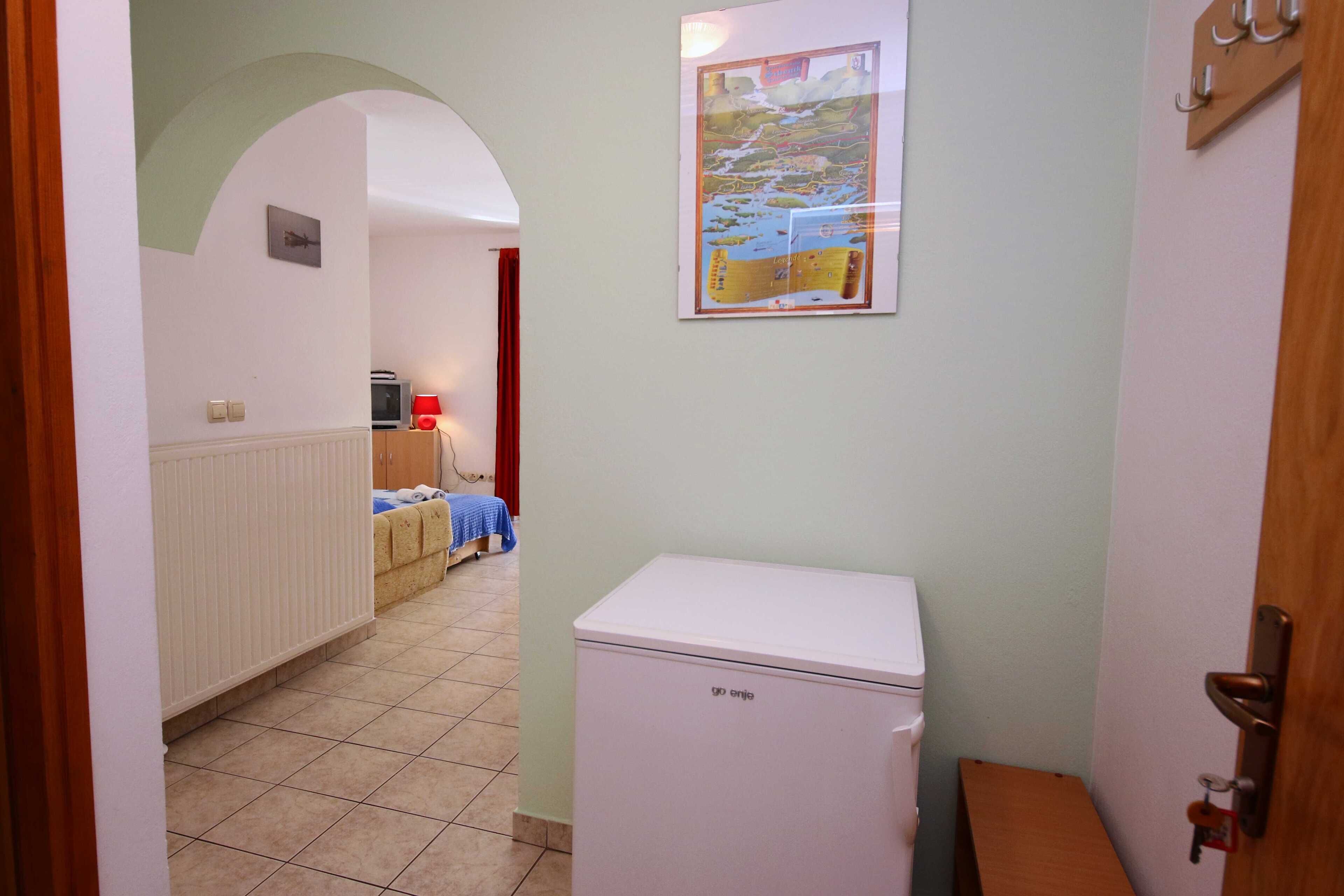 A narrow hallway with a white radiator, a small white refrigerator, and a bed with a blue and white striped bedspread.