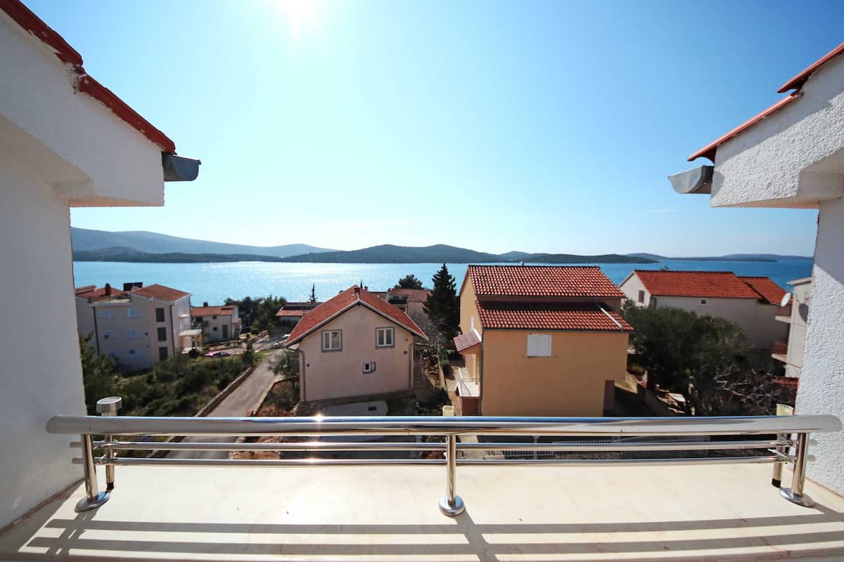 A balcony with a metal railing overlooking a residential area with houses and a body of water in the background.