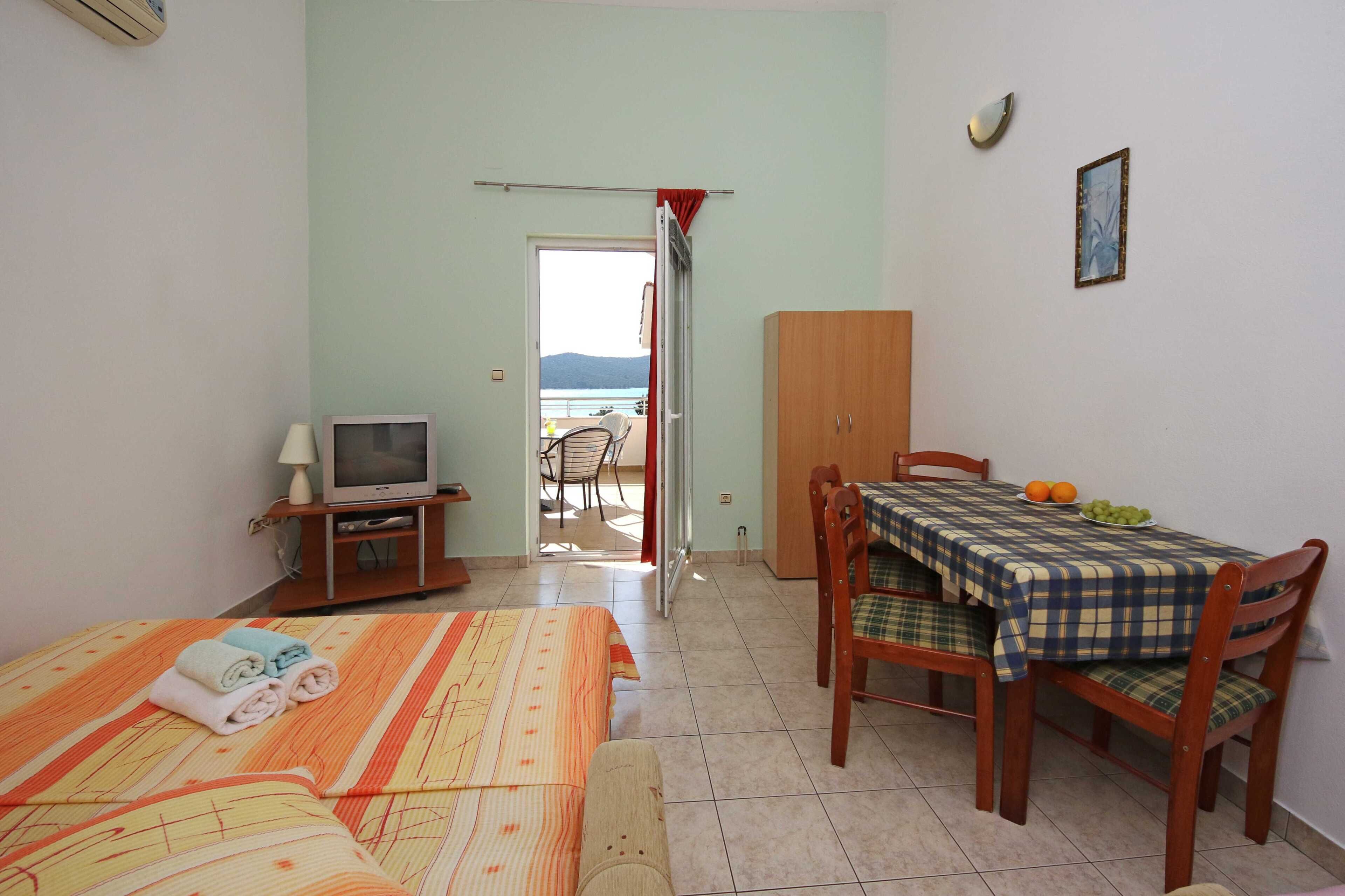 A room with a bed, a television, a dining table with chairs, and a view of the sea through an open door.
