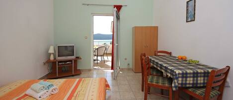 A room with a bed, a television, a dining table with chairs, and a view of the sea through an open door.