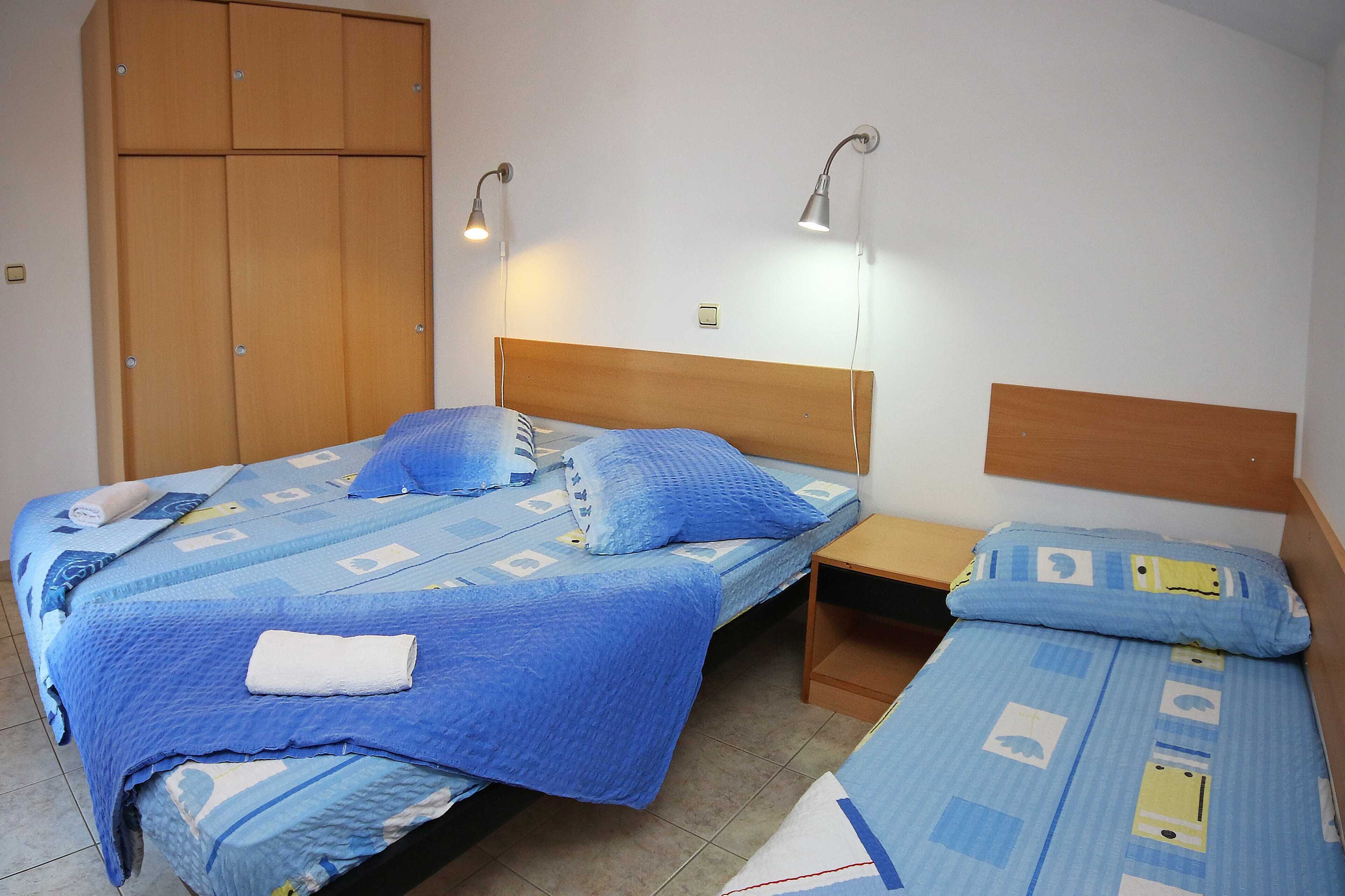 Uma cama de casal com colcha estampada em azul e branco, duas luminárias de cabeceira, um guarda-roupa de madeira e uma pequena mesa de cabeceira de madeira.