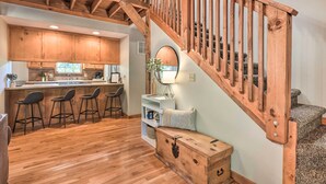 Una escalera de madera con barandilla, una zona de cocina con armarios de madera, una barra con taburetes y un pequeño rincón de estar con una lámpara.