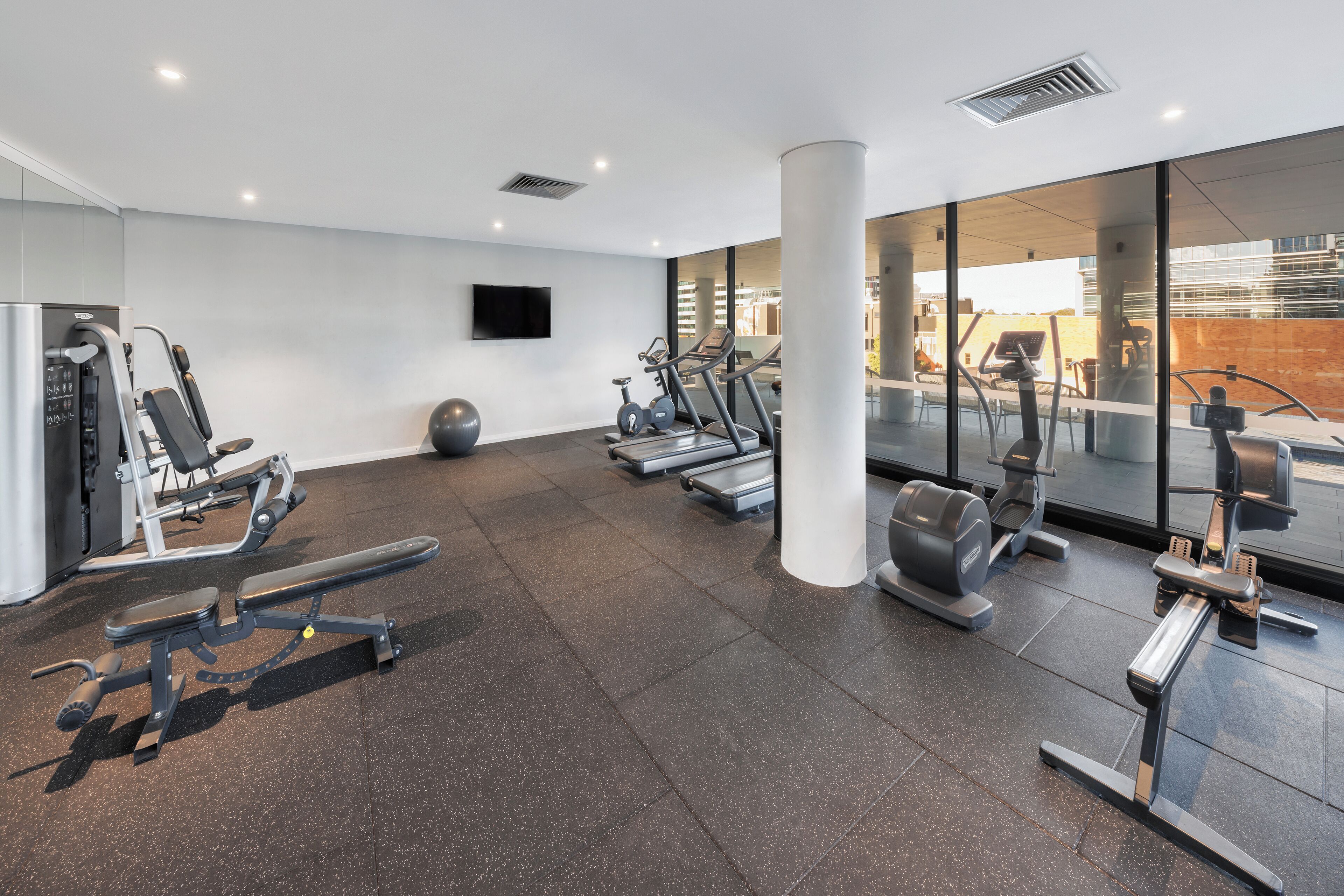 Un gimnasio moderno con varios equipos de ejercicio, un ventanal grande y un televisor de pantalla plana en la pared.
