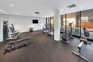 A modern gym with various exercise equipment, a large window, and a flat-screen TV on the wall. - Kooii Apartments (Fortitude Valley)