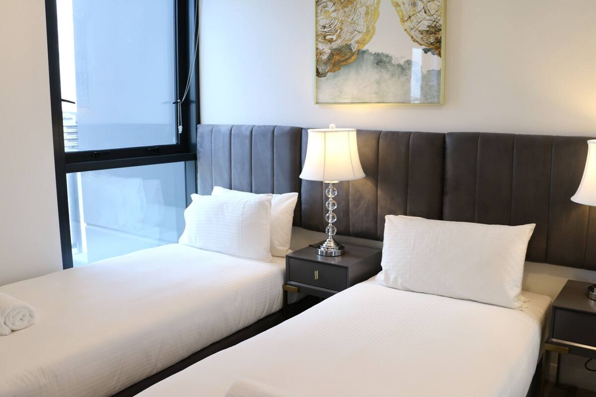 A hotel room with two beds, a nightstand with a lamp, a painting on the wall, and a window.