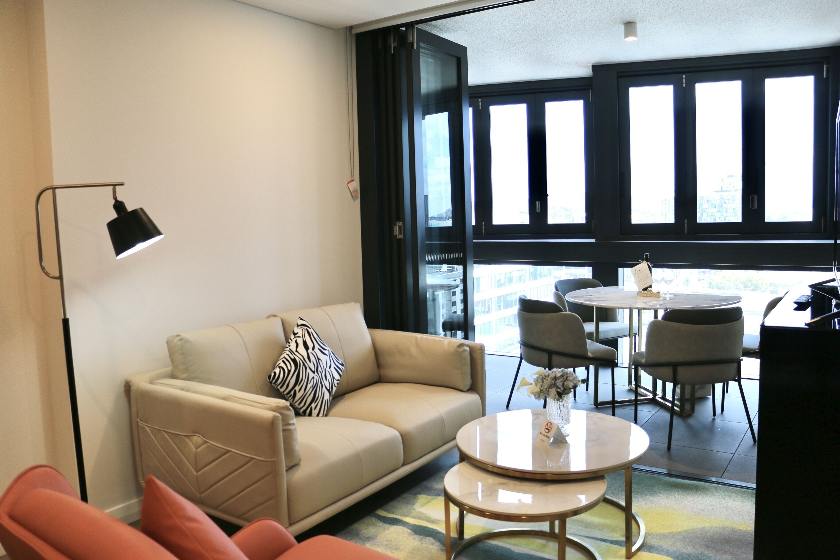 A modern living room with a beige sofa, a small round table, and a floor lamp. There is a dining area with a table and chairs visible through a glass door.