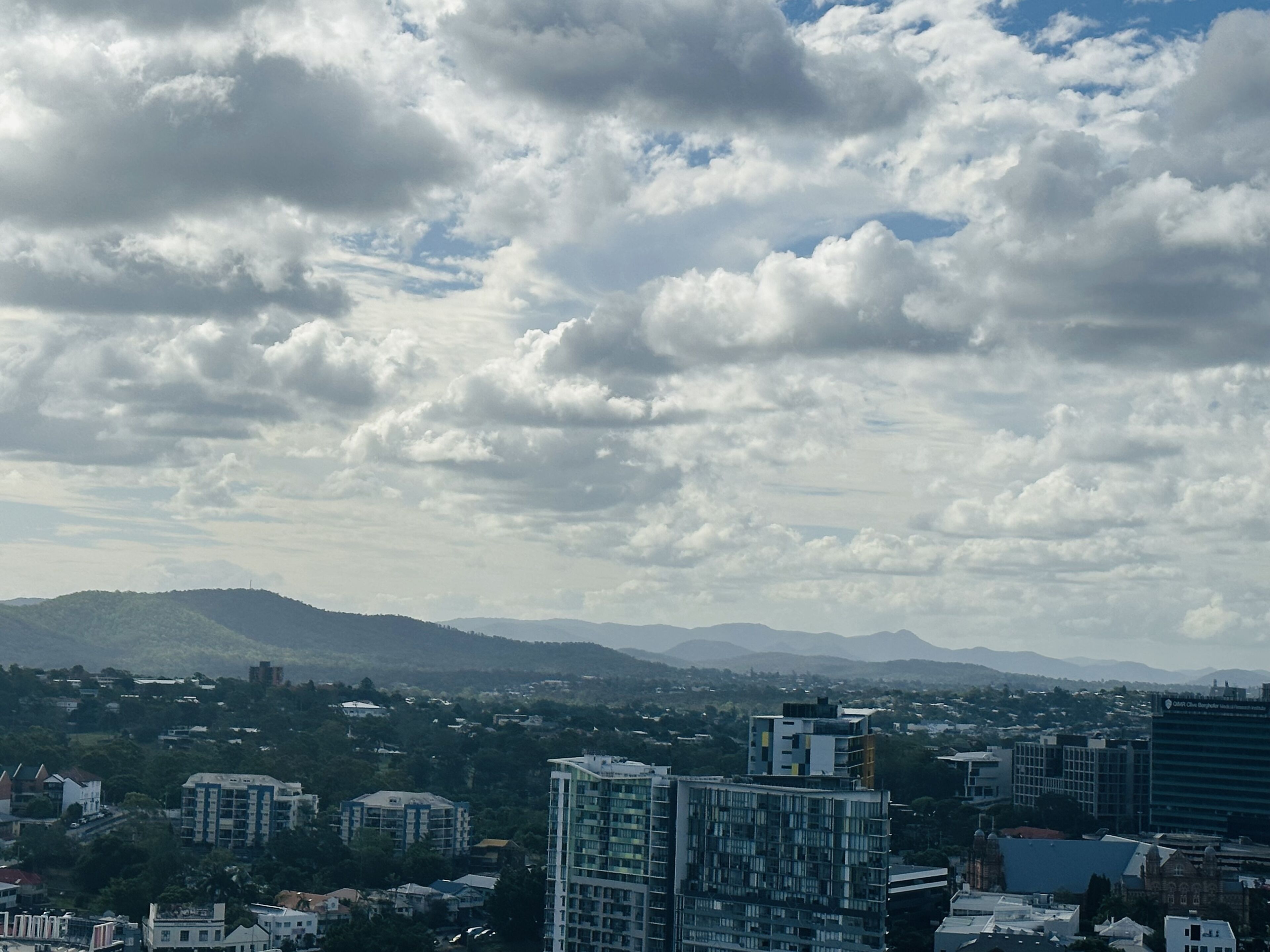 A cityscape with multiple high-rise buildings, a mountainous backdrop, and a partly cloudy sky.