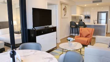 A modern hotel room with a flat-screen TV, a dining area with a small table and chairs, a sofa, and a kitchen area with a refrigerator and microwave.