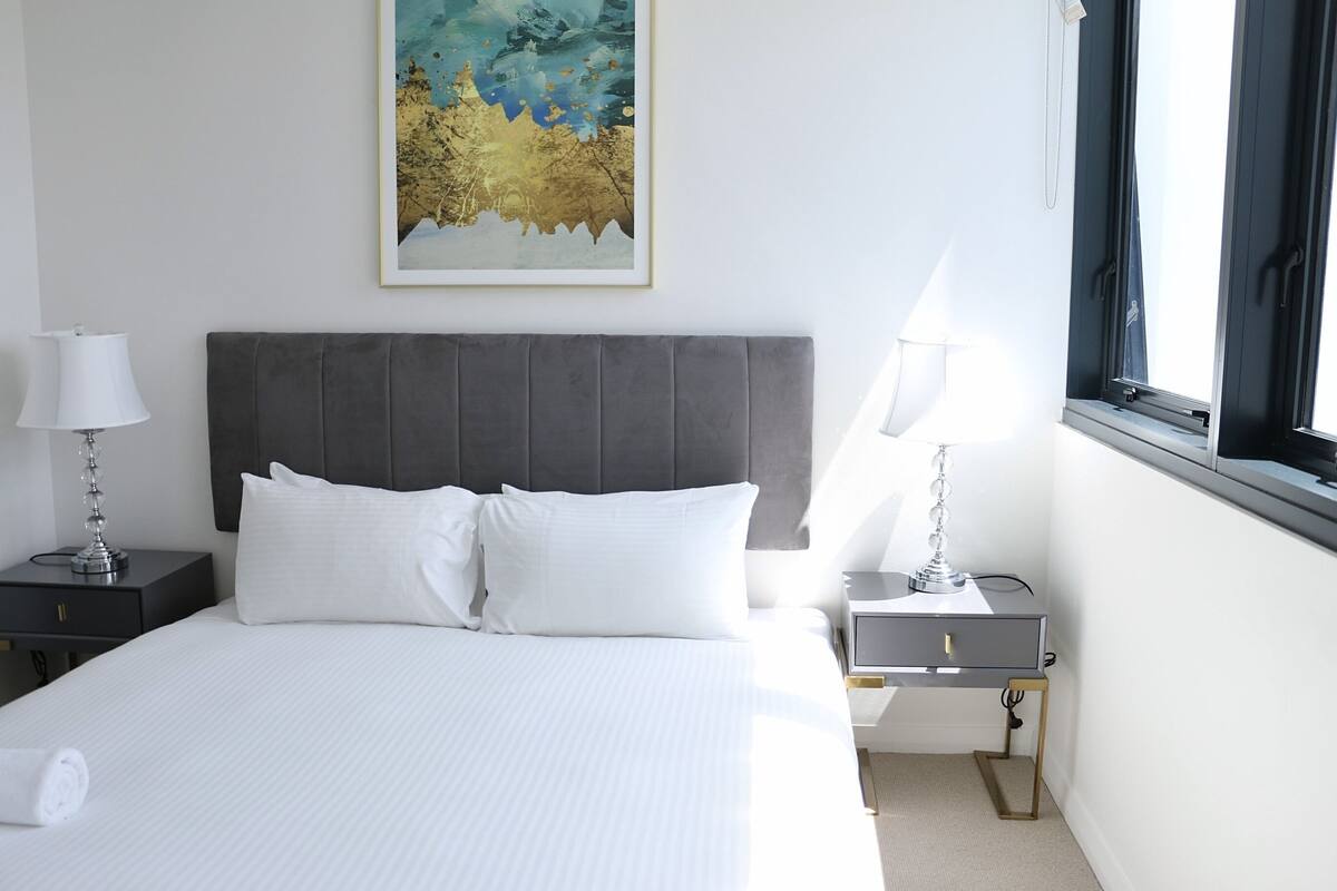 A neatly made bed with white linens, a grey upholstered headboard, and two bedside tables with lamps.