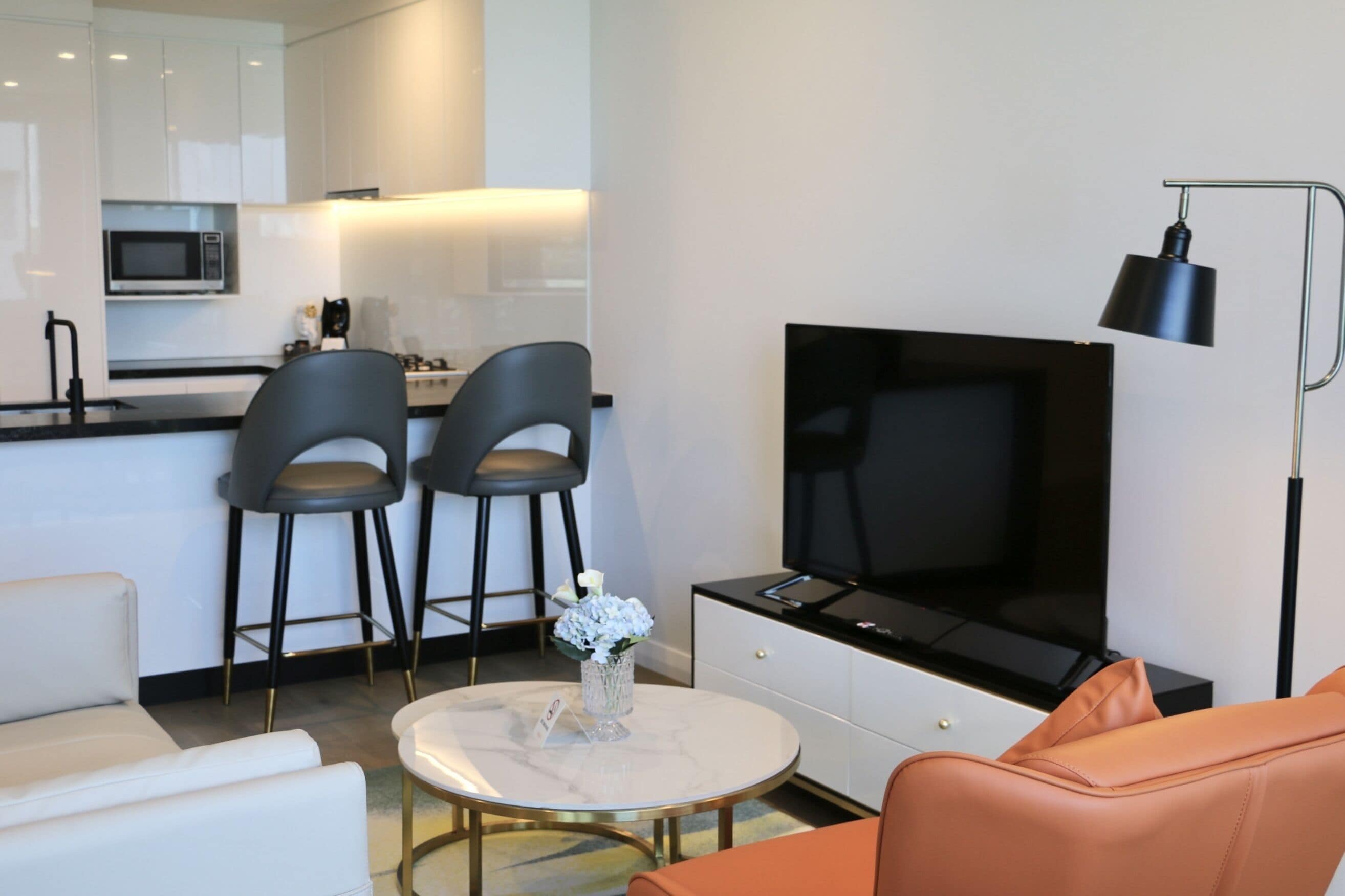 Ein modernes Wohnzimmer mit einer Couch, einem Couchtisch und einem Fernseher. Es gibt eine Küchenzeile mit einer Mikrowelle und einem Barhocker.
