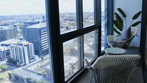 A high-rise room with a large window offering a city view, a wicker chair, and a small table. - Kooii Apartments (Fortitude Valley)