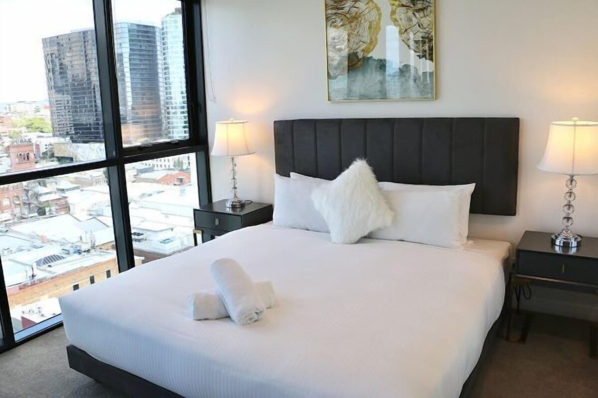 Una habitación de hotel con una cama grande, dos mesitas de noche, un ventanal con vistas a la ciudad y un cuadro en la pared.