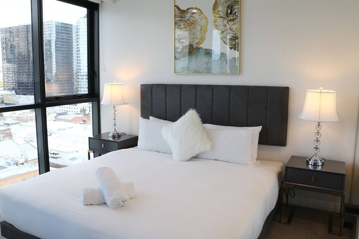 A hotel room with a large bed, two bedside tables with lamps, a view of the city through large windows, and a decorative wall piece above the bed.