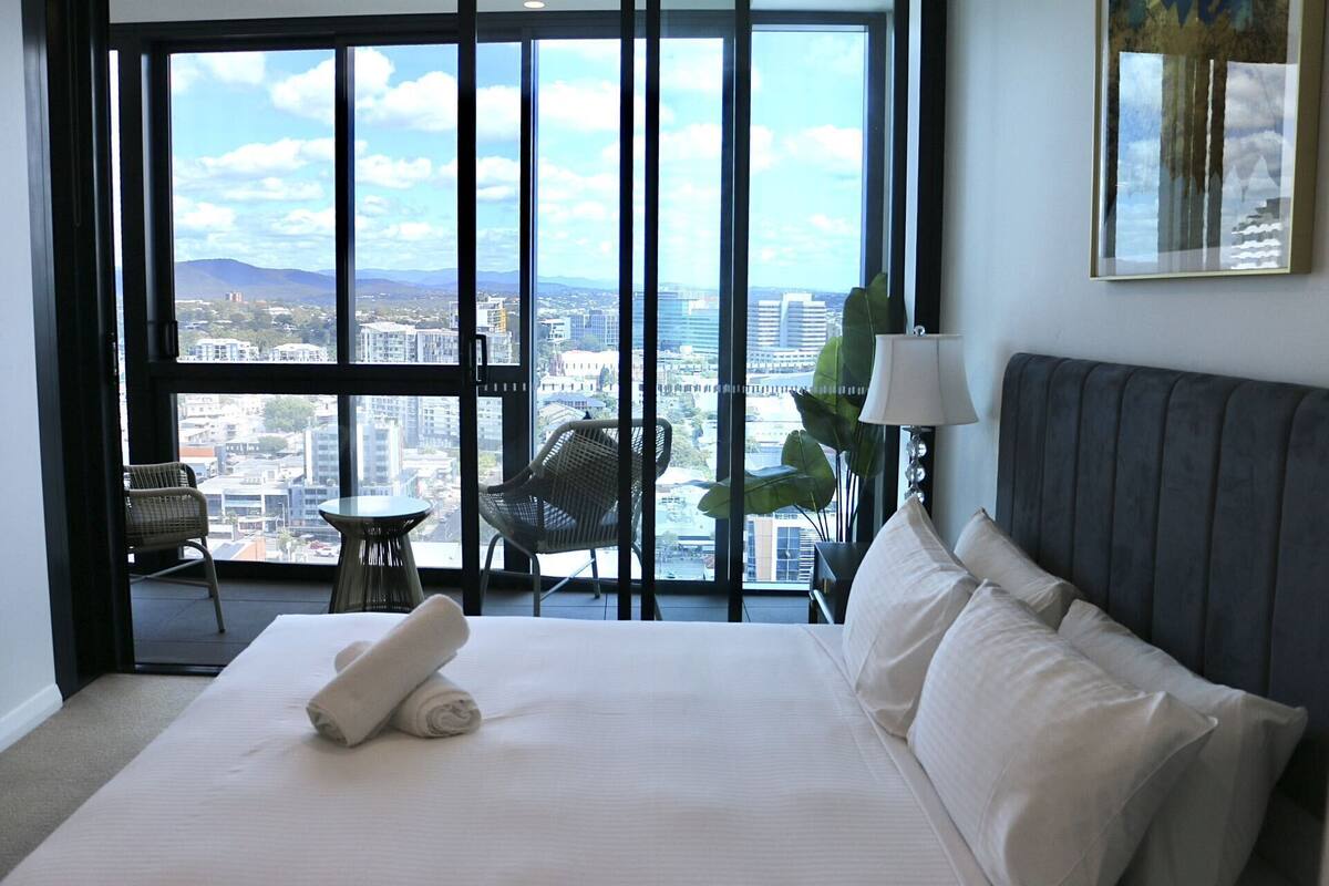 A hotel room with a large bed, a chair, a small table, and a view of the city through large windows.