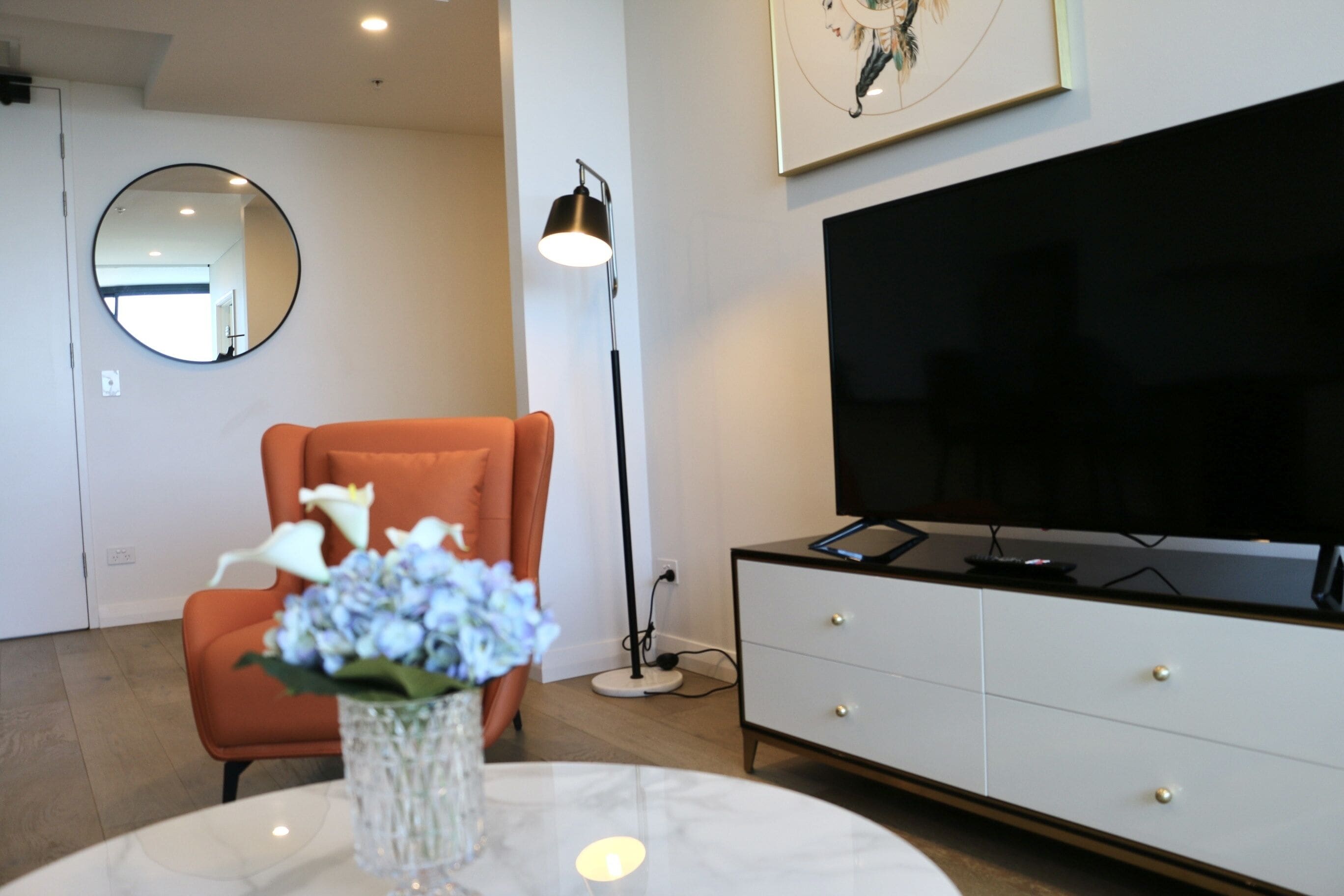 A modern living room with a round mirror, orange armchair, white cabinet, and a flat-screen TV.