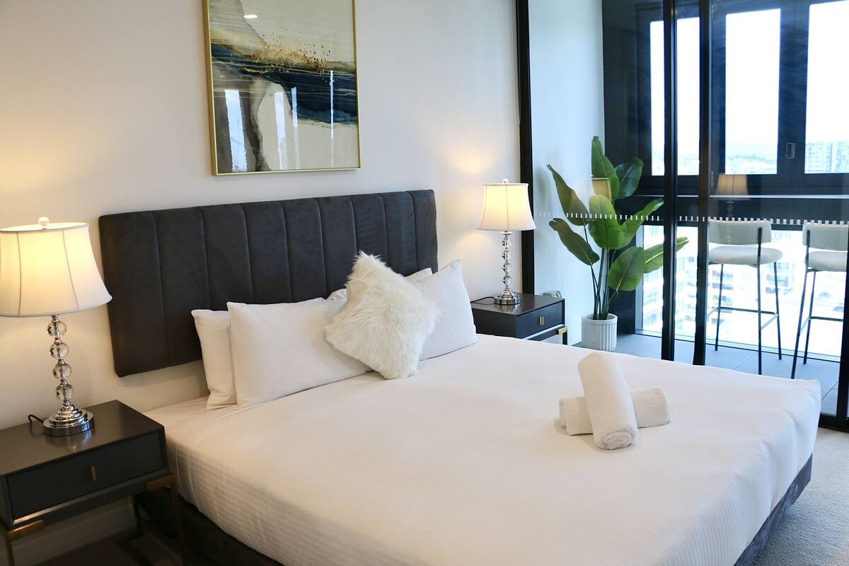 A neatly made bed with white linens and fluffy pillows, a bedside table with a lamp, and a large window with a view of the city.