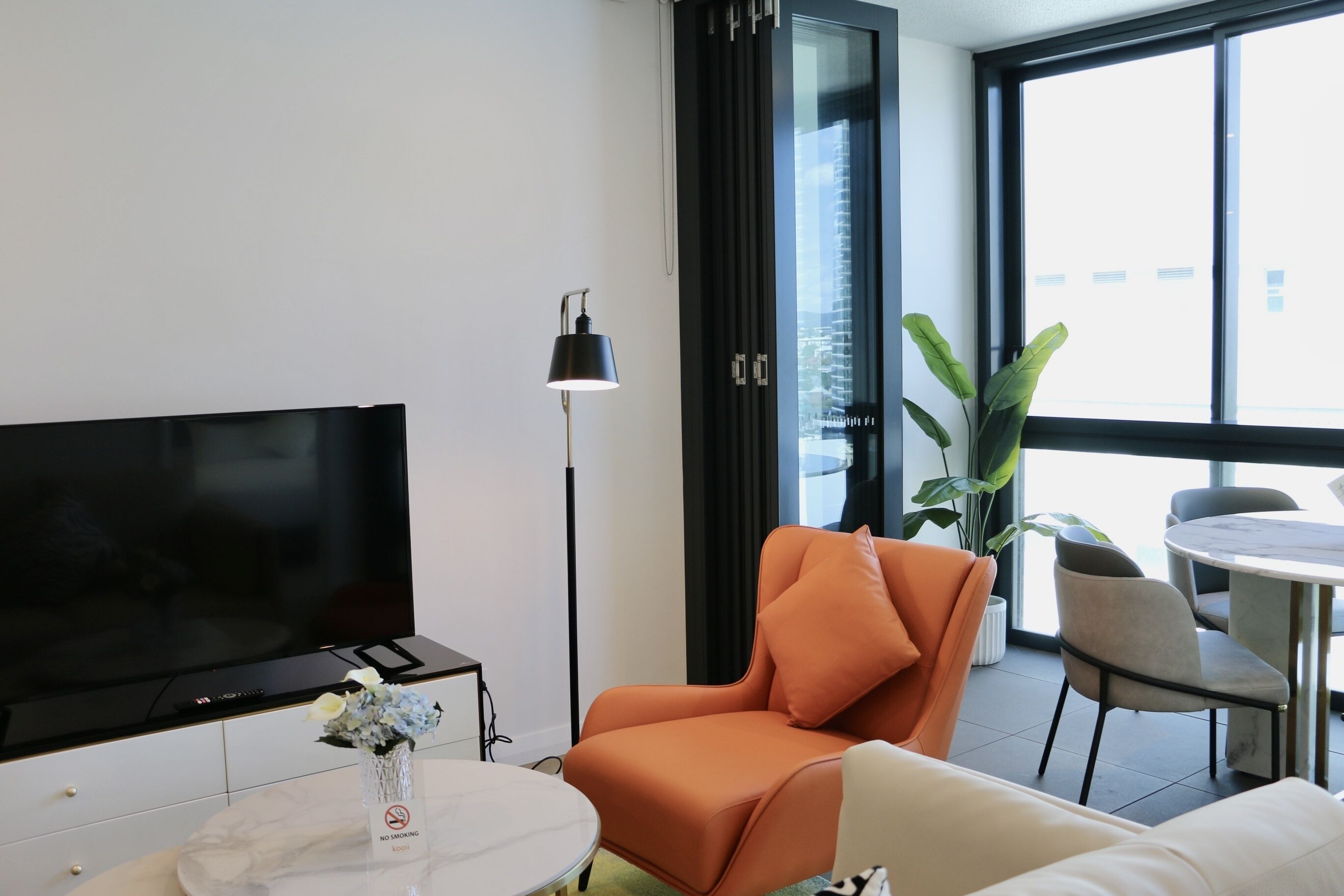 Ett modernt vardagsrum med en platt-TV, ett runt soffbord med en vas, en orange fåtölj, en vit soffa och en matplats med stolar och ett bord.