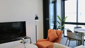 Ett modernt vardagsrum med en platt-TV, ett runt soffbord med en vas, en orange fåtölj, en vit soffa och en matplats med stolar och ett bord.