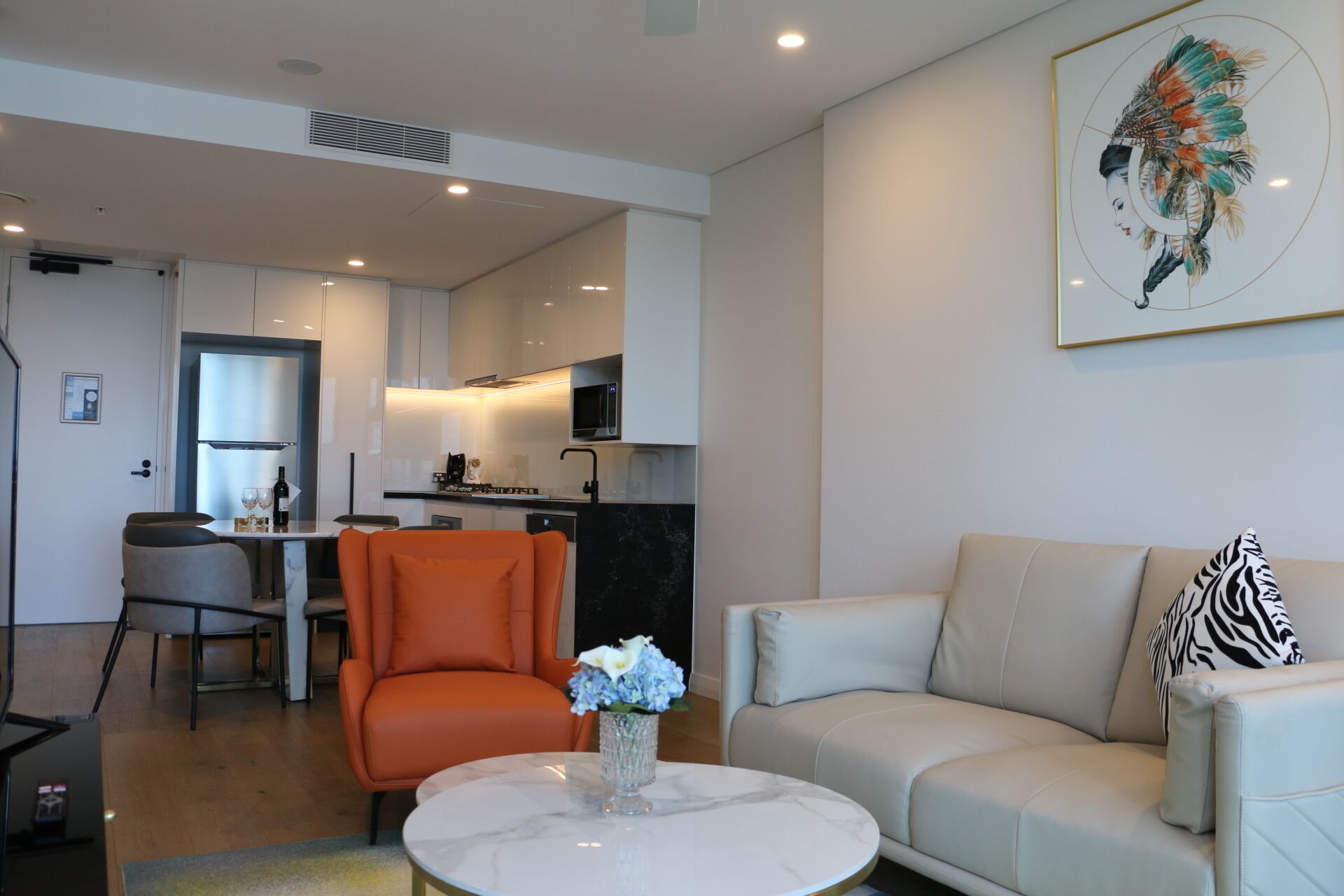 Een moderne woonkamer met een bank, een fauteuil en een salontafel. In de achtergrond is een keuken met een eettafel en stoelen.
