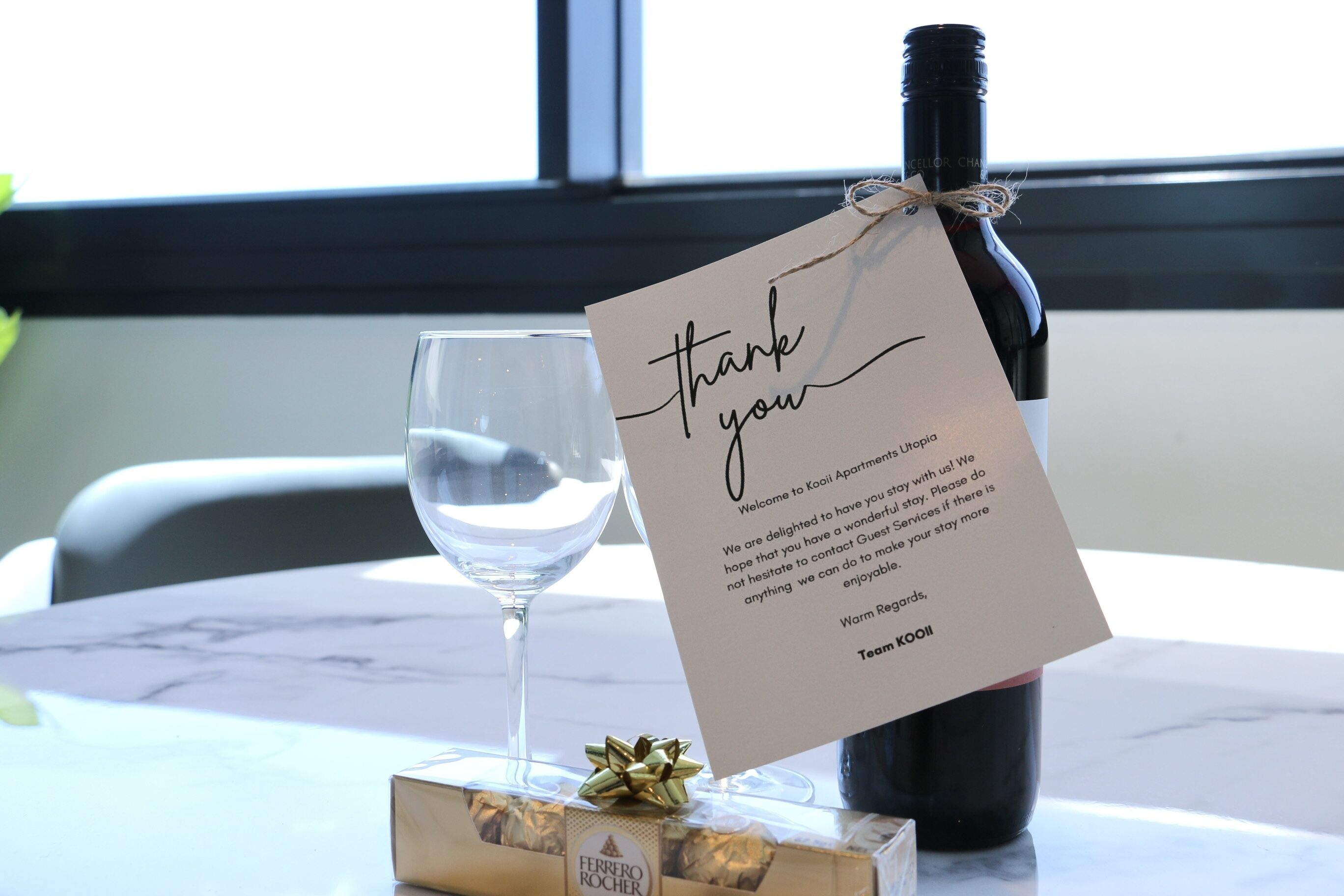 Una botella de vino, una copa, una tarjeta de agradecimiento y una caja de regalo sobre una mesa.