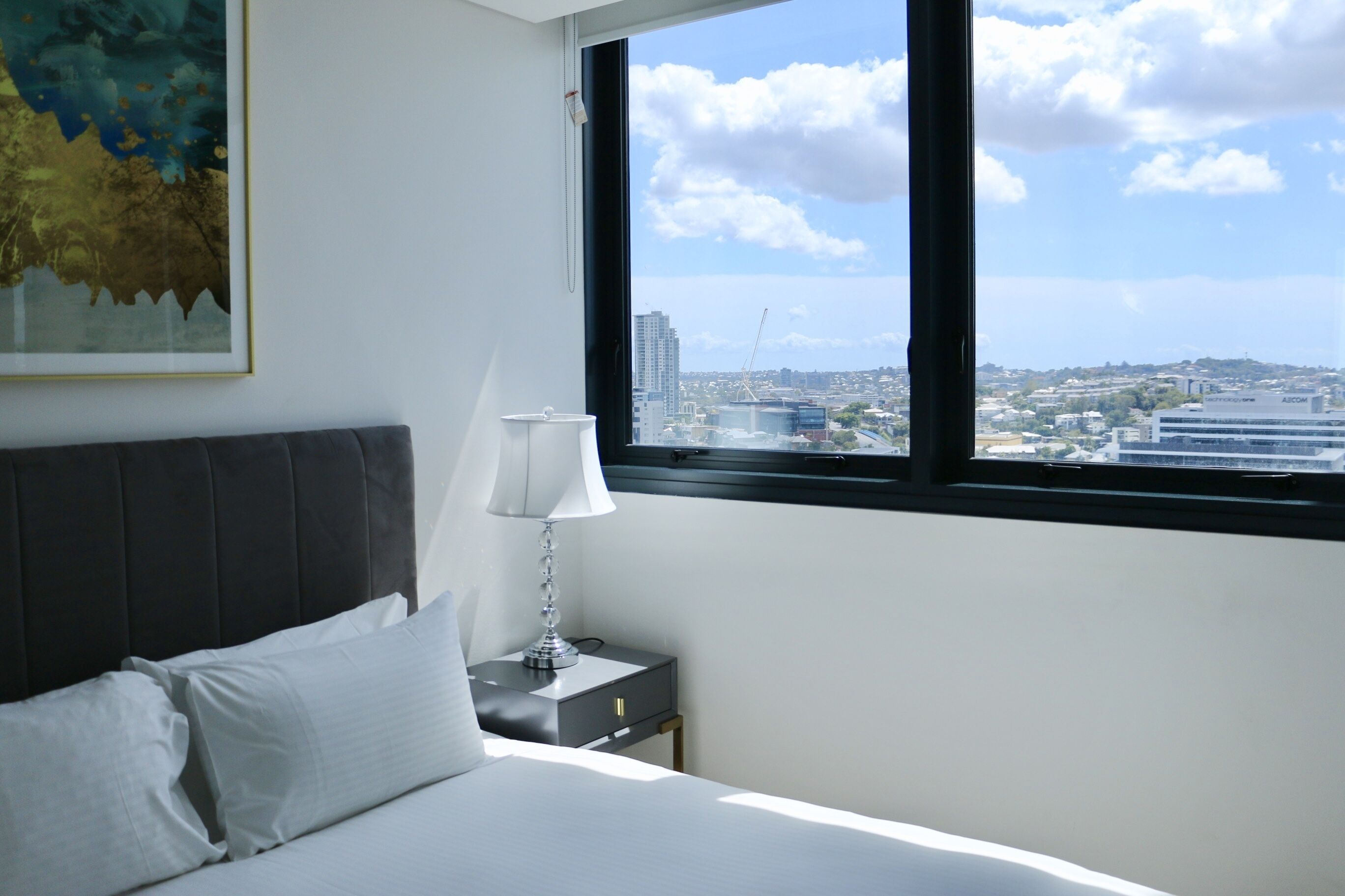 A bedroom with a bed, a nightstand, a lamp, and a window offering a city view.