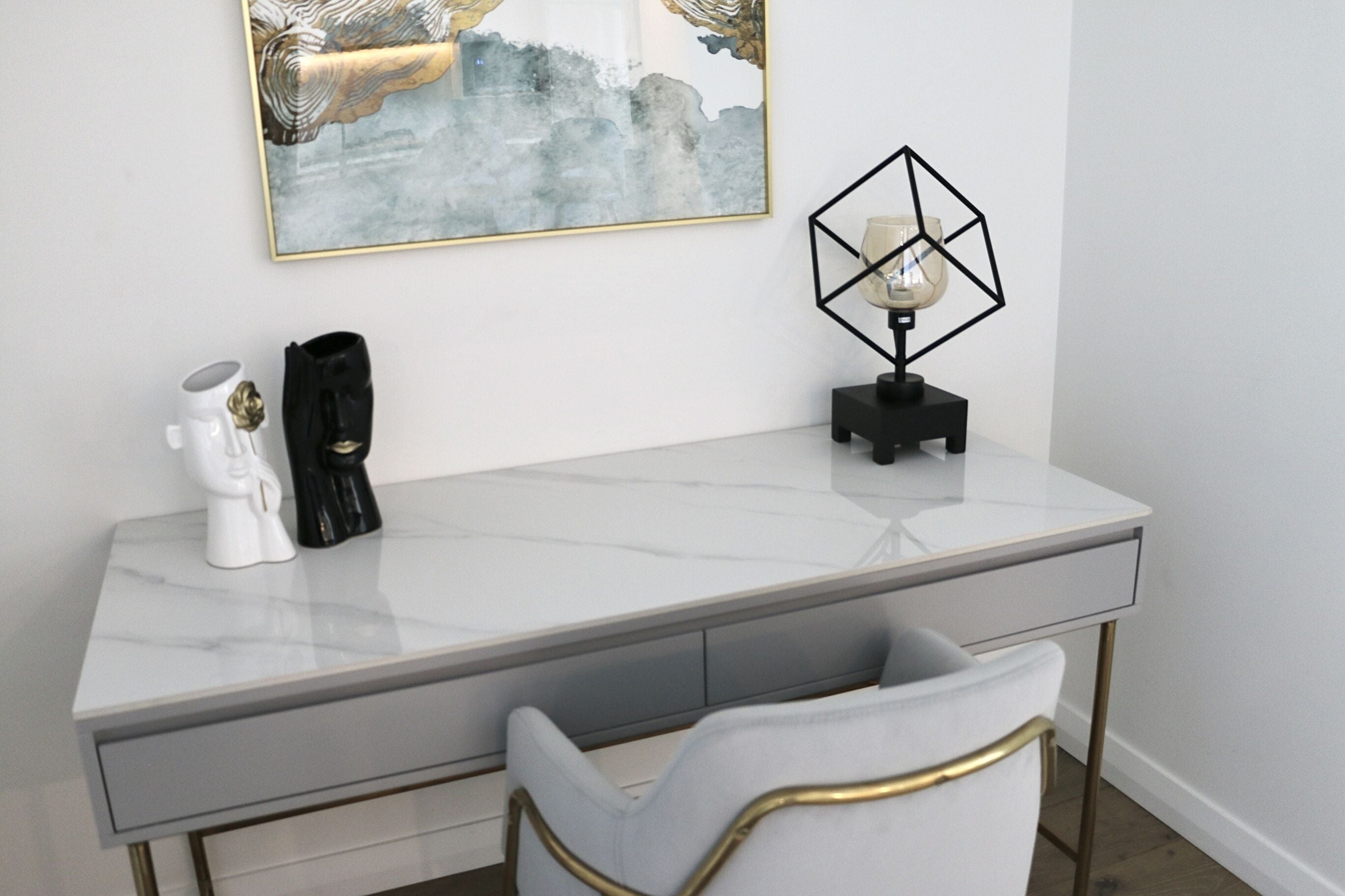 Ett modernt skrivbord med en bordsskiva i marmor, en vit stol med guldfärgade detaljer, en dekorativ vas och en lampa med geometriskt mönster.