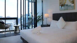 A hotel room with a large bed, a nightstand, a lamp, a chair, and a view of the city through floor-to-ceiling windows. - Kooii Apartments (Fortitude Valley)