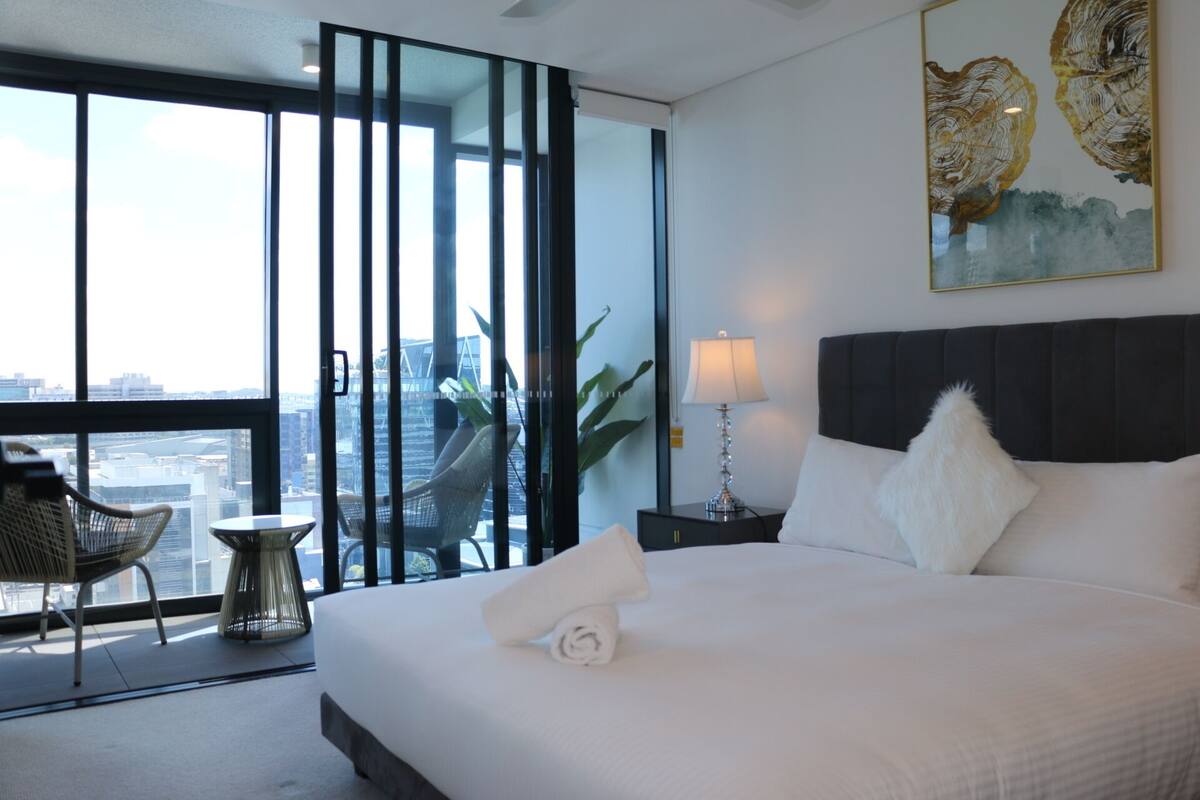 A hotel room with a large bed, a nightstand, a lamp, a chair, and a view of the city through floor-to-ceiling windows.
