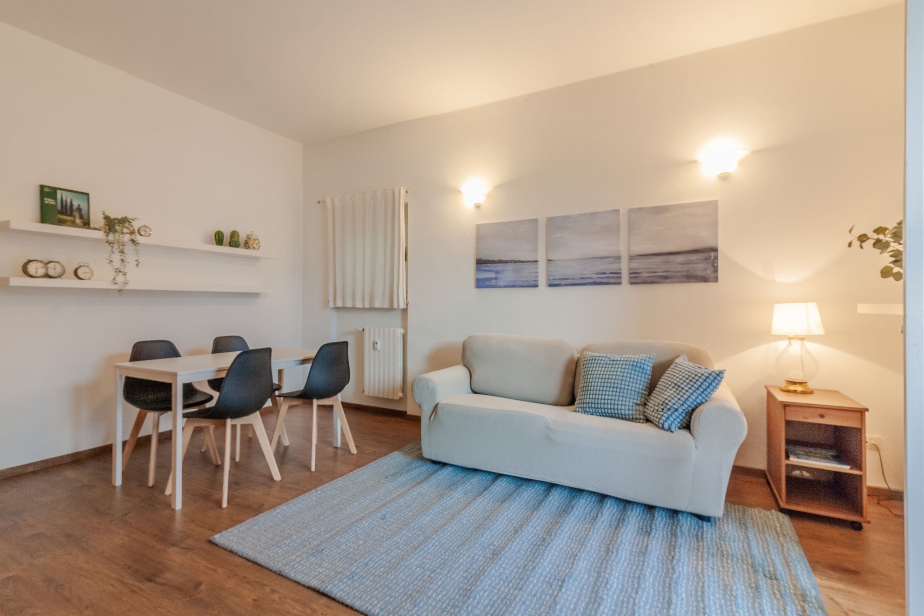 A modern living room with a white sofa, a dining table with chairs, a blue rug, and wall-mounted shelves with decorative items.