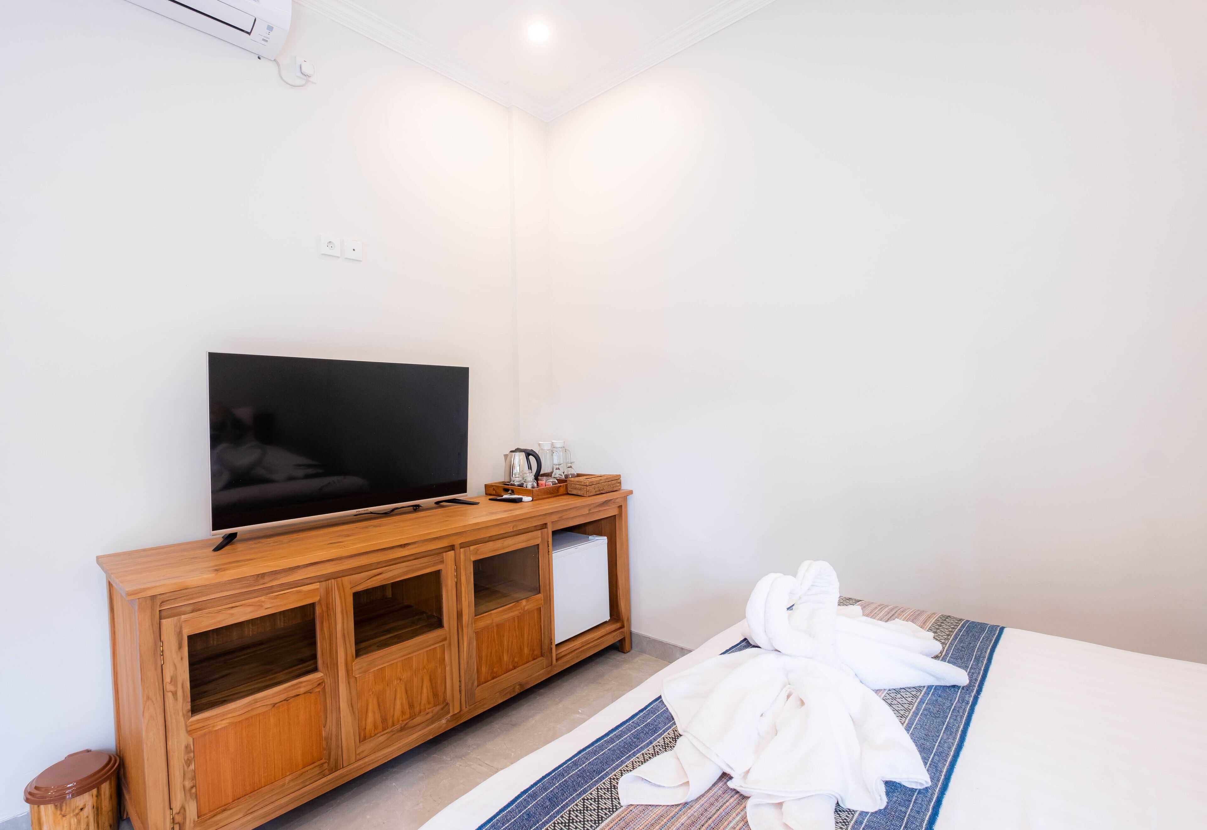A hotel room with a wooden TV stand, a flat-screen TV, a small white refrigerator, and a neatly made bed with a patterned bedspread.