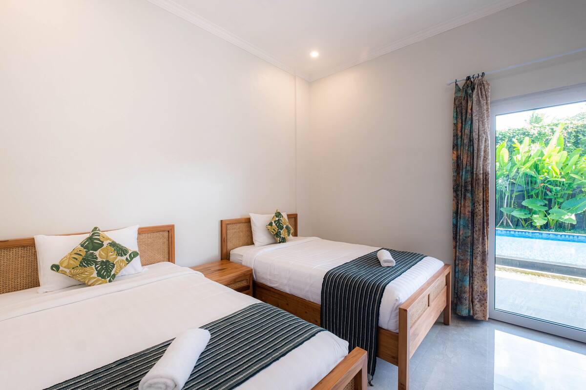 A hotel room with two beds, a window with curtains, and a view of greenery outside.