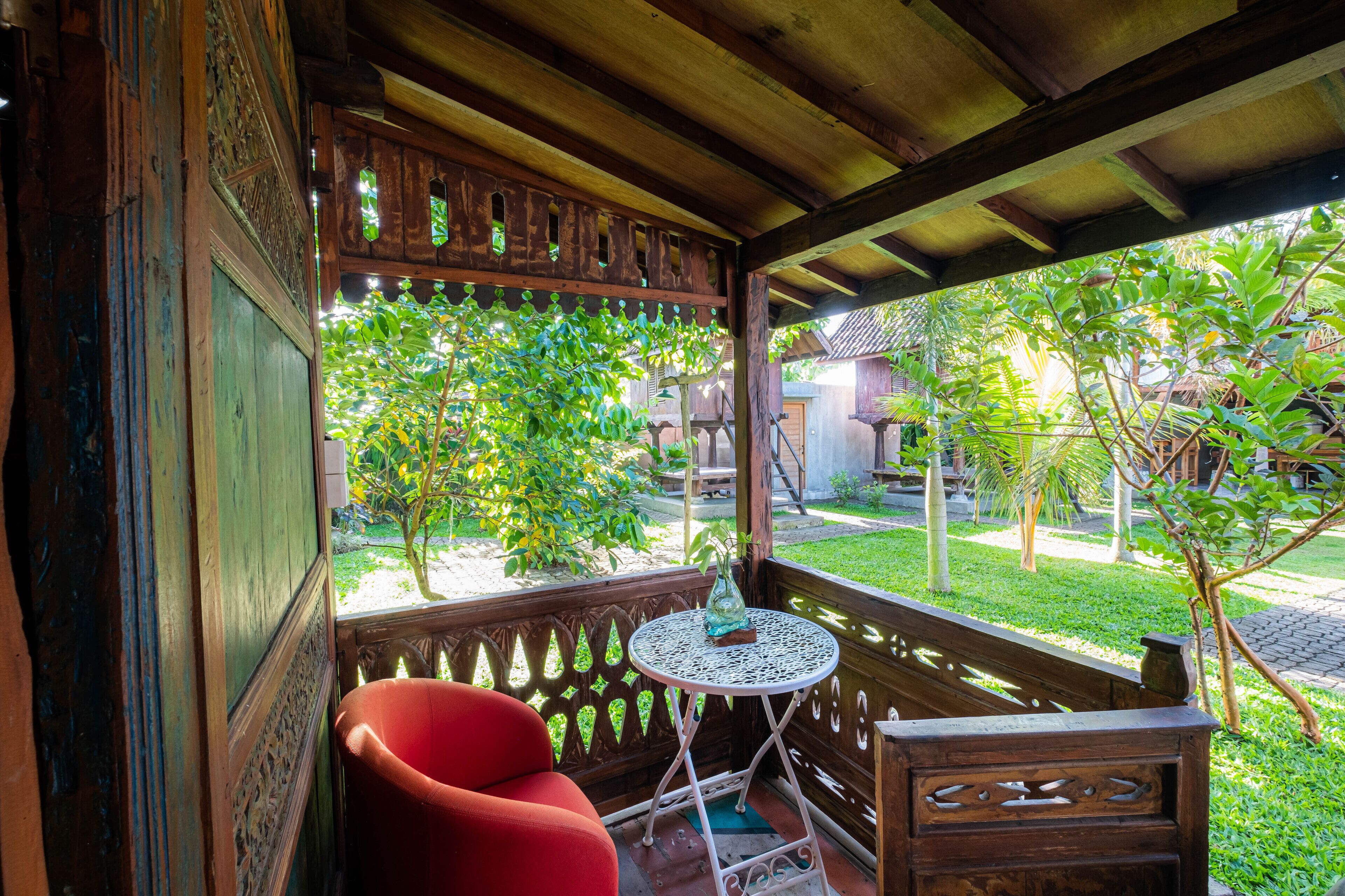 Une véranda en bois avec une chaise rouge et une petite table, donnant sur un jardin avec des arbres et un bâtiment en arrière-plan.