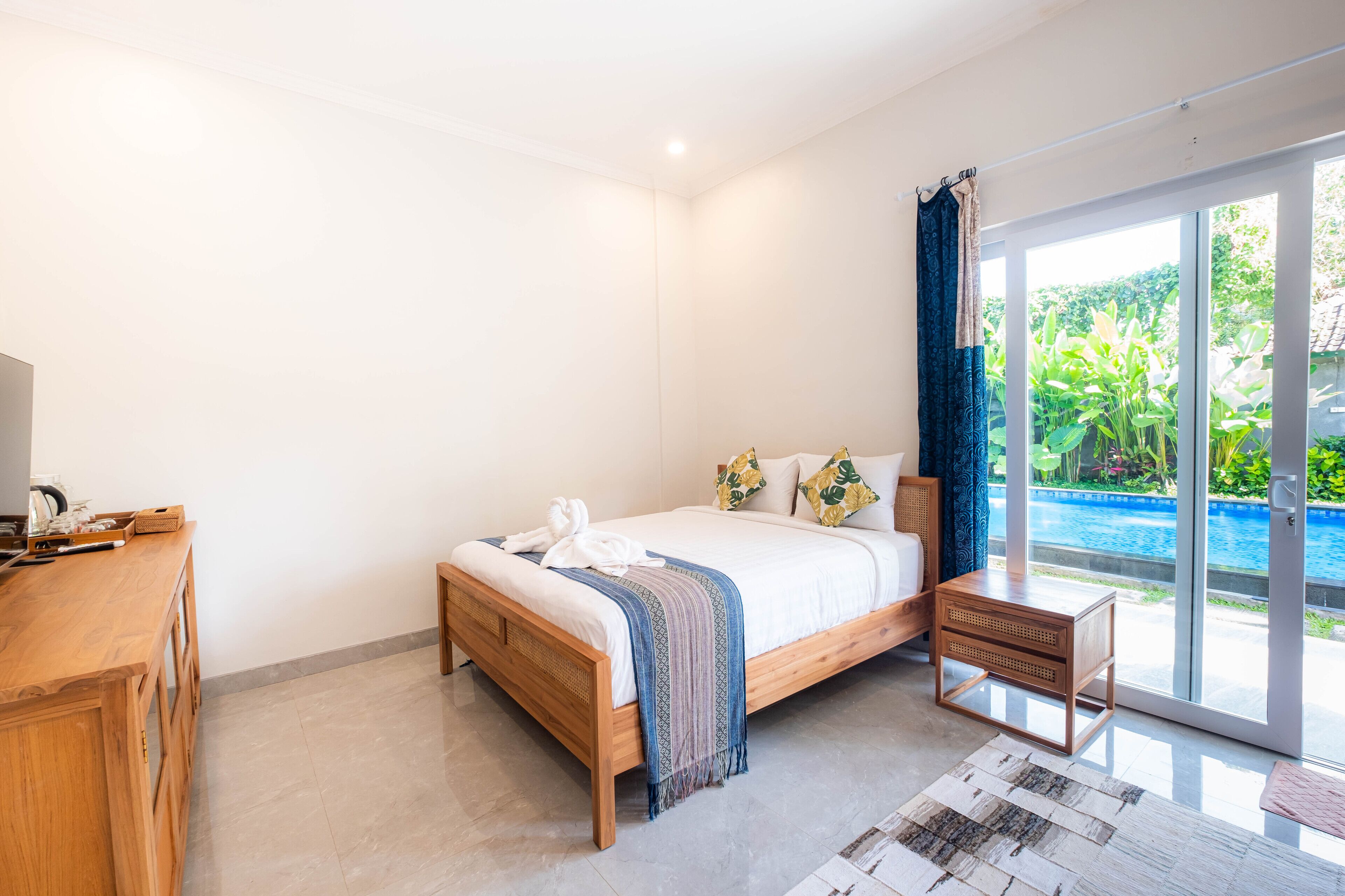 A bedroom with a wooden bed, a small bedside table, a TV, and a view of a swimming pool through a glass door.