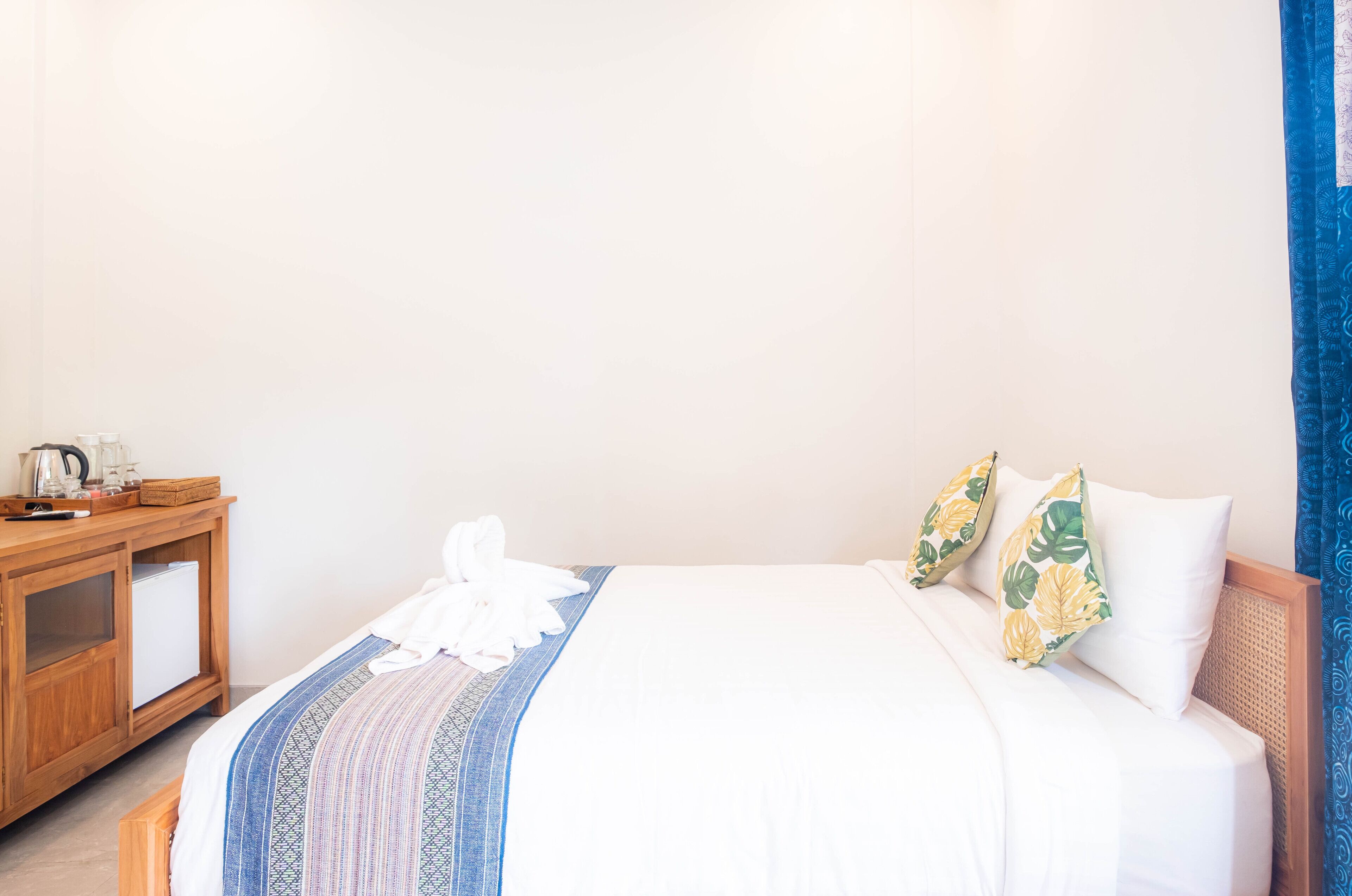 A neatly made bed with a patterned bedspread, a bedside table with a kettle and a basket, and a blue curtain.