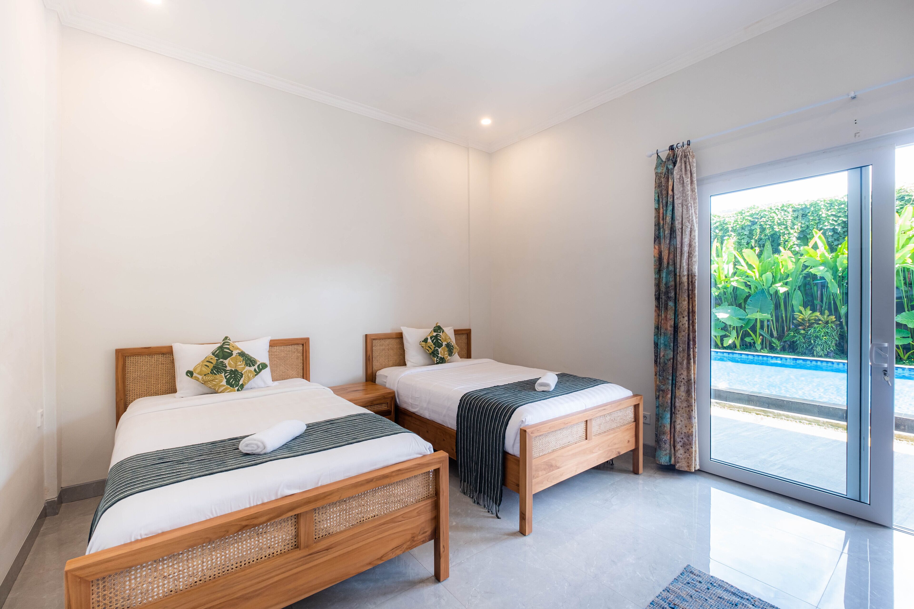 A hotel room with two wooden beds, a large sliding glass door to a balcony, and a view of greenery outside.