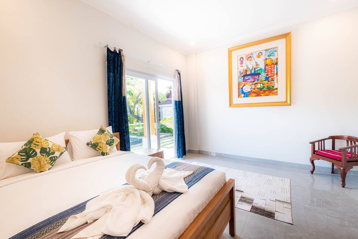 A hotel room with a bed, a wooden chair, a painting on the wall, and a view of the outdoors.