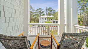 Un balcon avec des fauteuils en osier, une petite table et une vue sur une maison blanche entourée d’arbres.