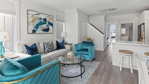 Una sala moderna con un sofá, sillones color turquesa, una mesita de centro y una escalera que lleva a una cocina.