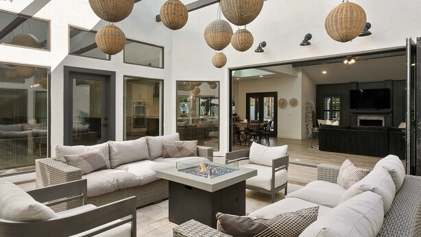 A spacious outdoor living area with a sectional sofa, a coffee table, and wicker furniture. There are hanging wicker lamps, a dining area, and a kitchen visible through glass doors. - SWSS 6BR Luxury Group Retreat | Chef's Kitchen, Pool, Pickleball & Putting Green (Scottsdale)