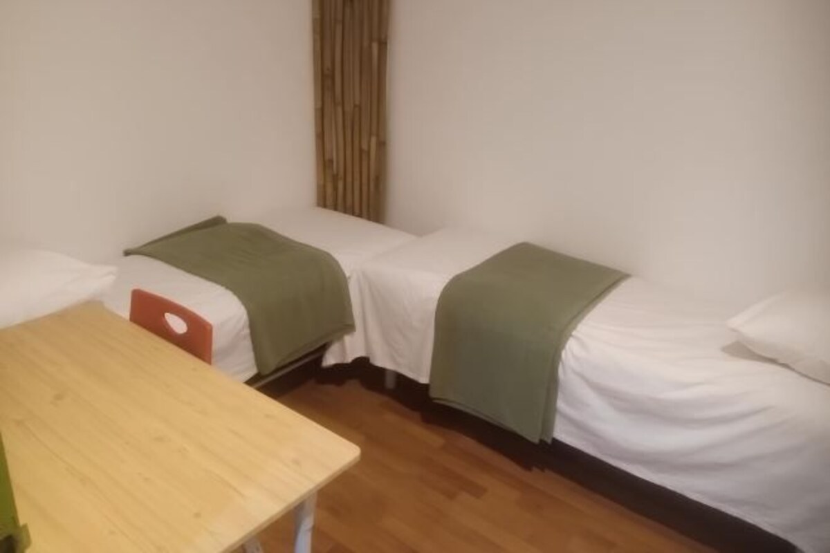 A small, clean room with two single beds, a wooden table, and a bamboo curtain.