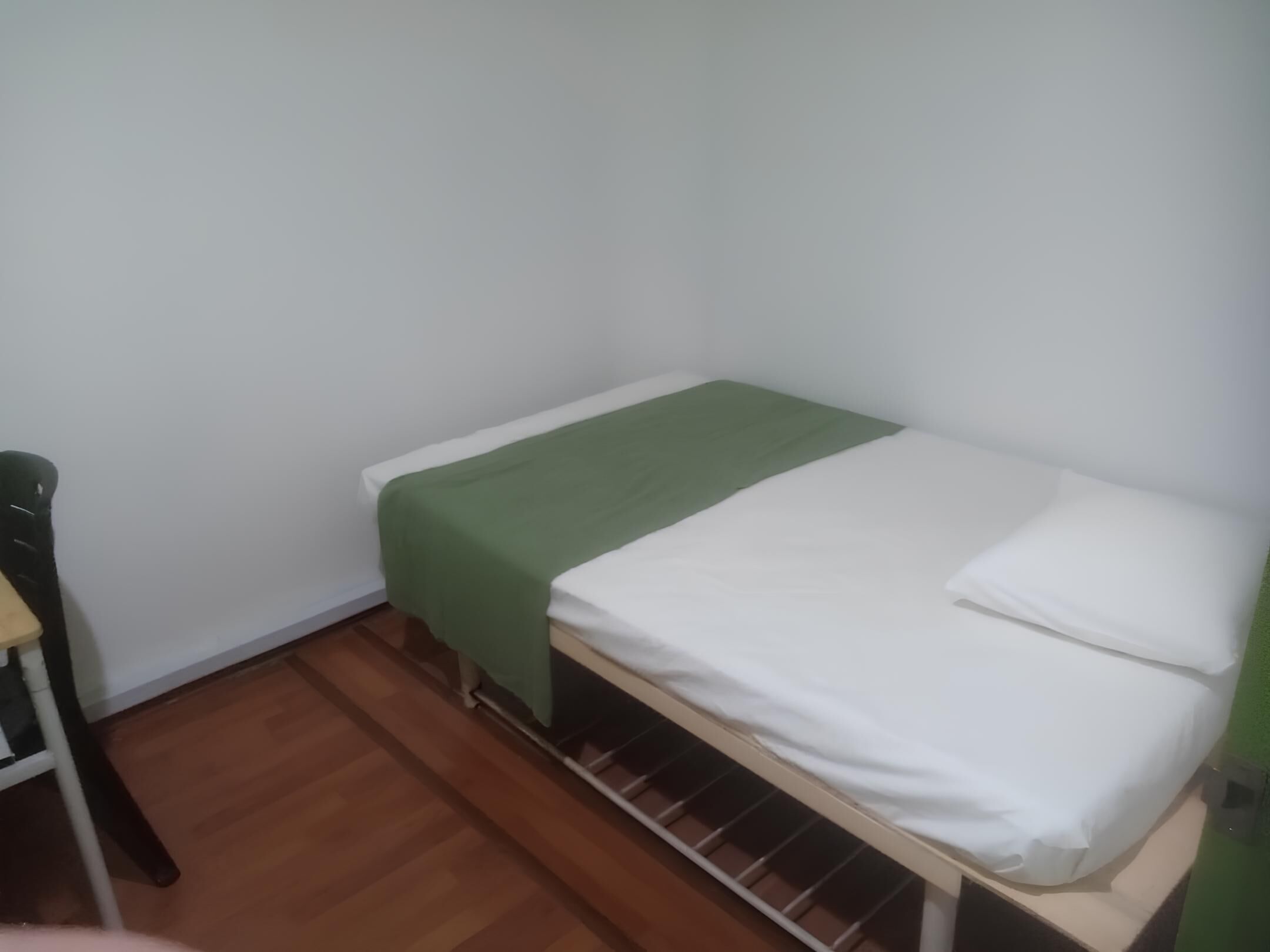 Una habitación pequeña con cama individual, colcha verde, ropa blanca y suelo de madera.