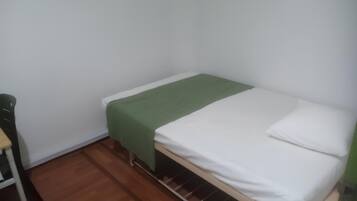 Una habitación pequeña con cama individual, colcha verde, ropa blanca y suelo de madera.