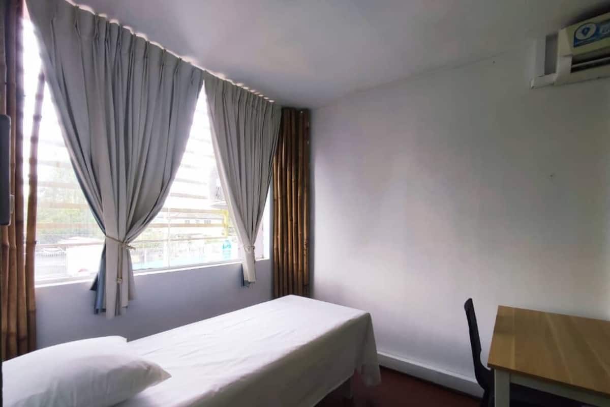 A single-bed room with a wooden table and a chair, a window with curtains, and a white wall.