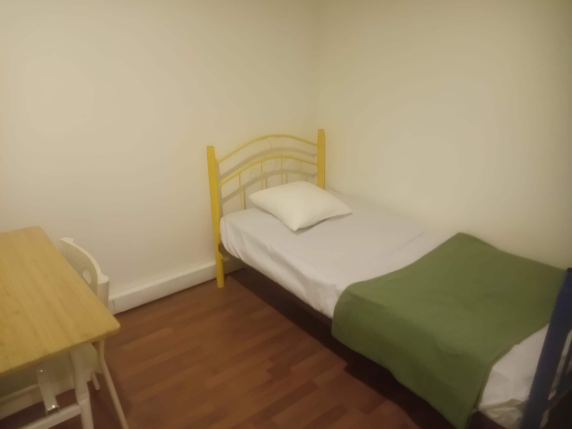 Una habitación pequeña con cama individual, piso de madera, estructura de cama de metal amarillo, almohada blanca y manta verde.
