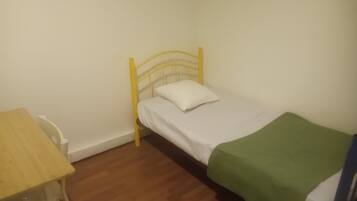 Una habitación pequeña con cama individual, piso de madera, estructura de cama de metal amarillo, almohada blanca y manta verde.