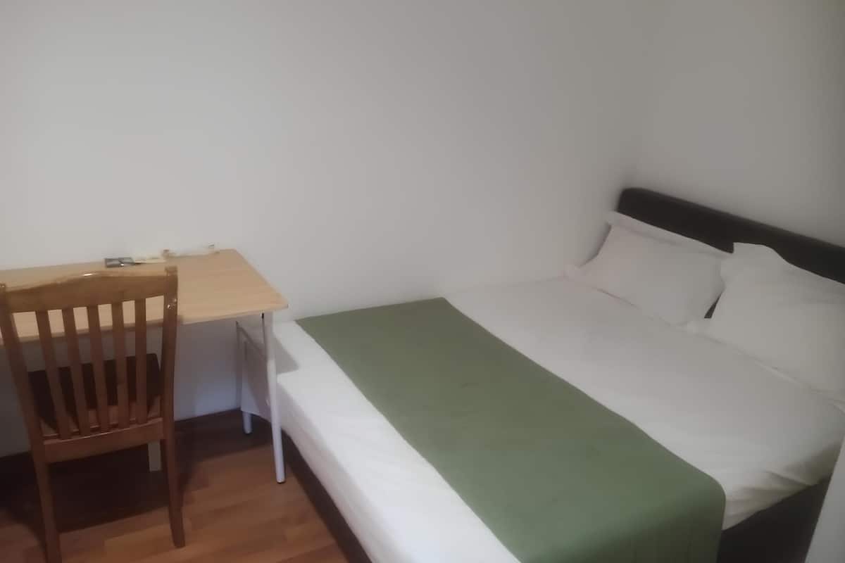 A small, simple room with a wooden desk and chair, a single bed with a green blanket, and white bedding.