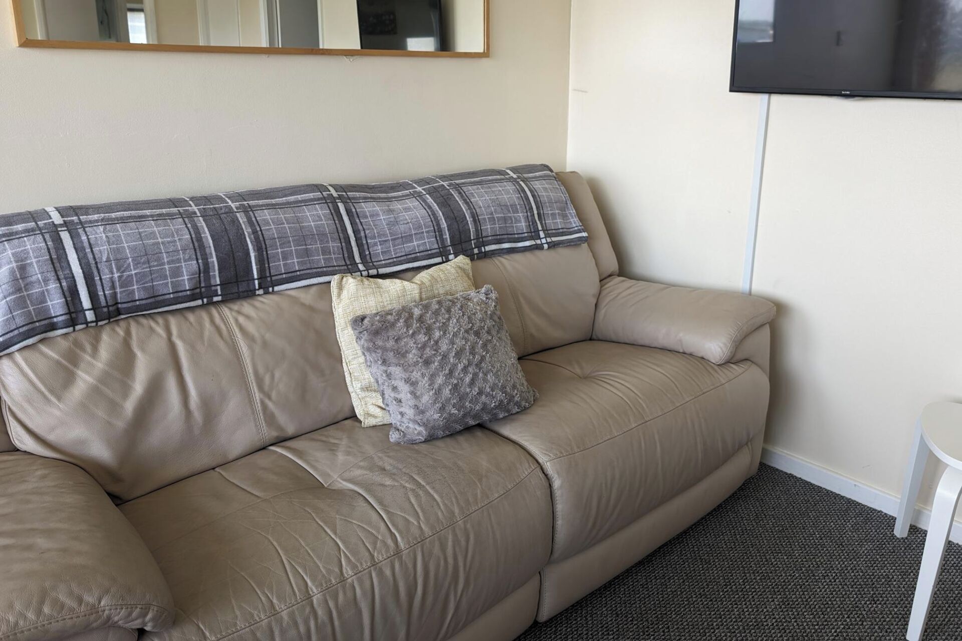 En beige lädersoffa med en rutig kudde och en fluffig grå kudde, ett litet vitt bord och en väggmonterad platt-TV.