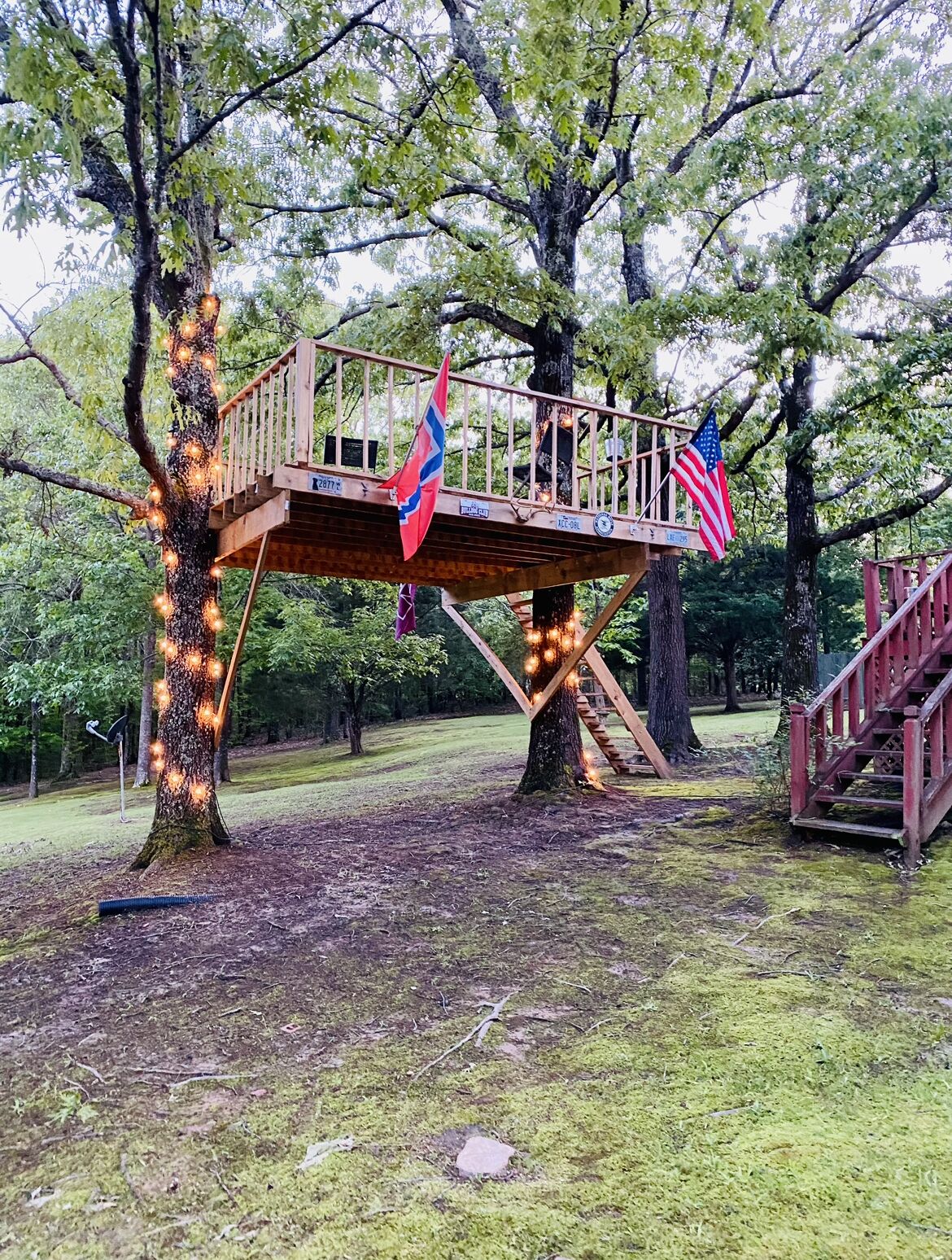 Une terrasse en bois, ornée de deux drapeaux, entourée d’arbres et bordée d’une pelouse.